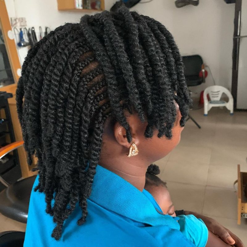 Pretty woman with natural kinky twists that are also very thick sits in a salon chair holding a baby