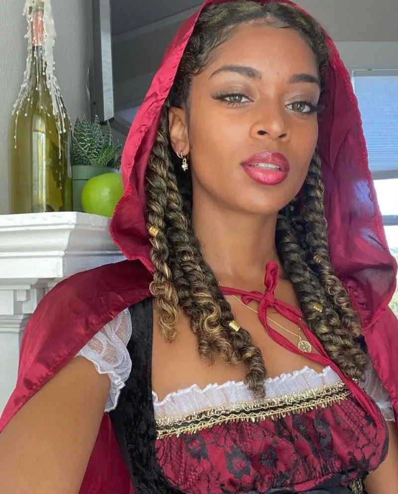 Woman with loose natural kinky twists with a red riding hood-inspired outfit with the hood up stands in front of a mantle