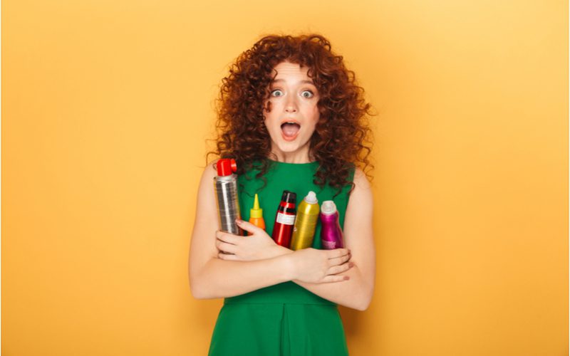 Portrait of a curly haired woman in a green dress holding an armful of hair products standing in an orange room