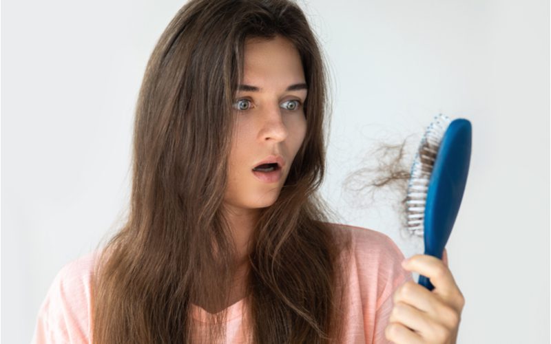 For a piece on how to stop hair shedding, a woman looking in disbelief at her purple brush that has multiple clumps of hair in it