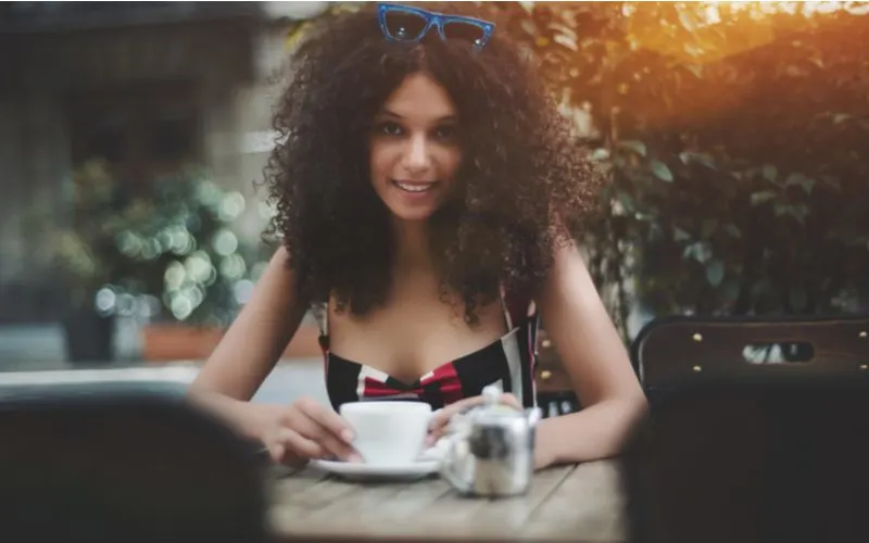 Pretty lightskin woman in a patterned dress with coily hair and glasses on her head holds an espresso glass