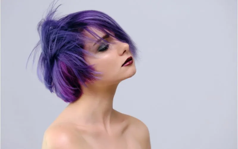 Woman with purple feathered hair wearing red lip and purple eyeshadow and no shirt stands in a lilac room letting a breeze blow