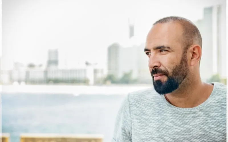 Spanish man with a buzzcut and a beard grins standing in front of a bay with a skyline in the background