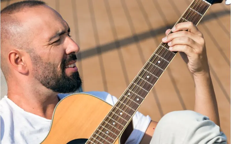 Man playing a guitar with a buzzcut hairstyle smiling and squinting his eyes