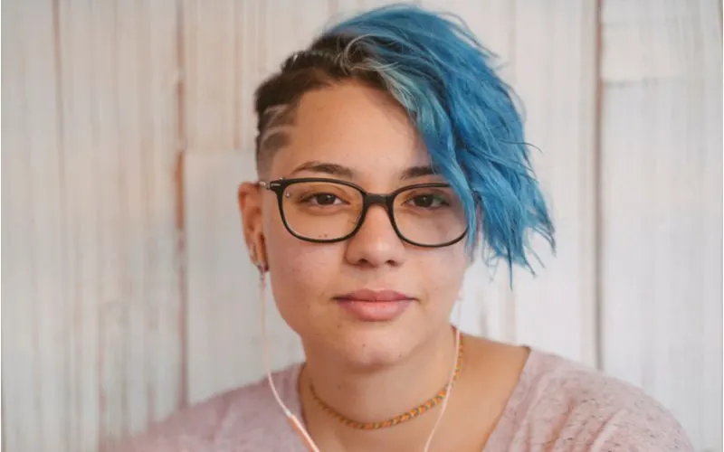 Liberal lesbian woman with blue hair and glasses with a shaved side rocking the punk hairstyle and listening to music on gold headphones