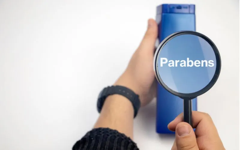 Woman holding a shampoo bottle that says Parabens under a magnifying glass to symbolize the second step in getting rid of greasy hair