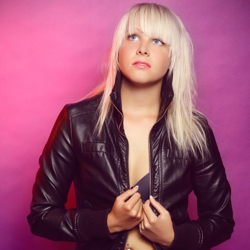 Woman with a blonde rocker shag haircut stands in a purple bra below a black leather jacket