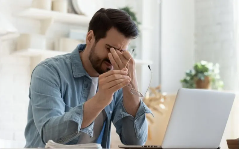 Man stressed out and holding his eyes with his glasses in his other hand with an open laptop symbolizing one of the ways to get rid of dandruff (mitigating stress)