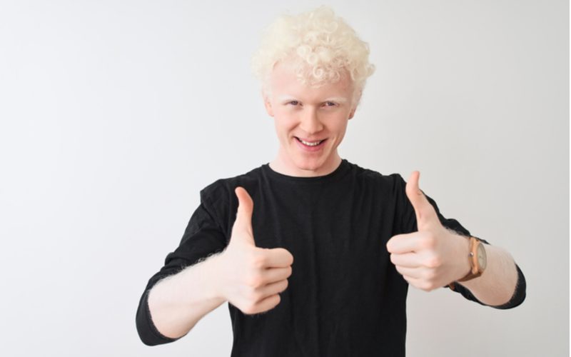 Pale man with bleached blonde hair wears a black shirt and gives a thumbs up
