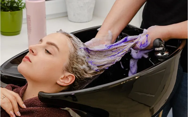 For a piece on what does purple shampoo do to brown hair, a woman with brown hair gets her hair washed with purple shampoo in a black sink