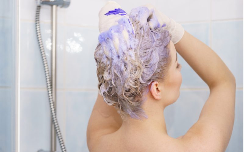 Brown-haired woman using purple shampoo on her hair in a home shower