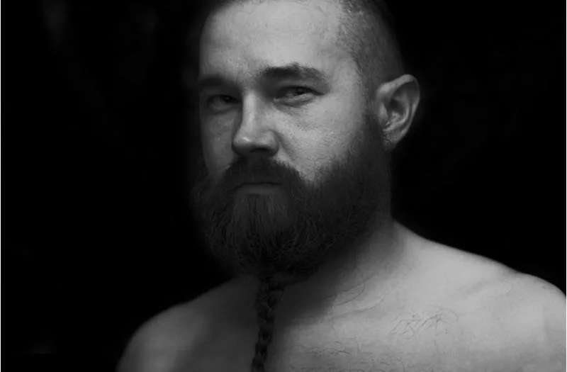 Man wearing a bearded braid while not wearing a shirt standing in a dark room