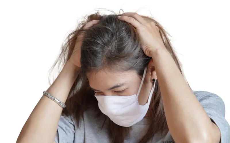 Woman holding her head because she's experiencing covid hair loss and wondering how to fix it while wearing a mask