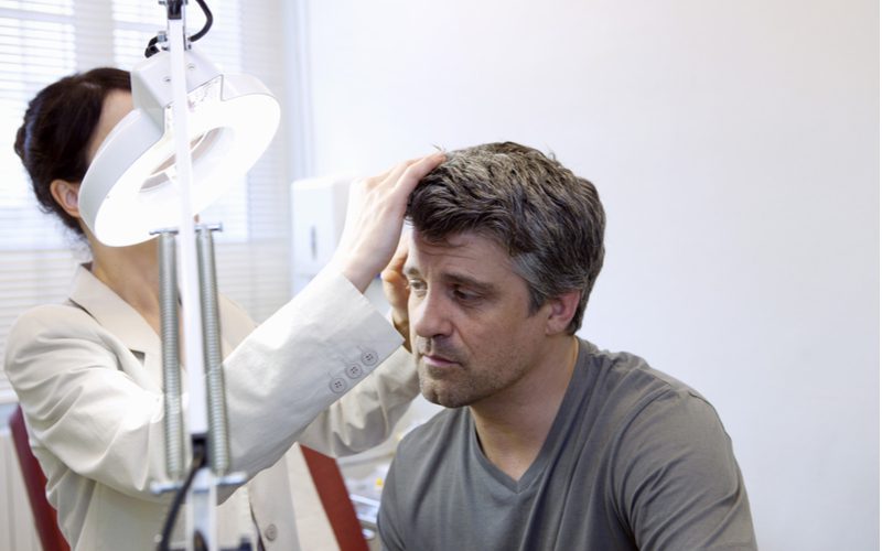 Image for a piece on how to get rid of dandruff featuring a dermatologist examining a man's scalp under a bright white light