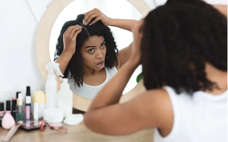 Woman looking at her scalp trying to figure out why her hair hurts while standing wearing a white tank top