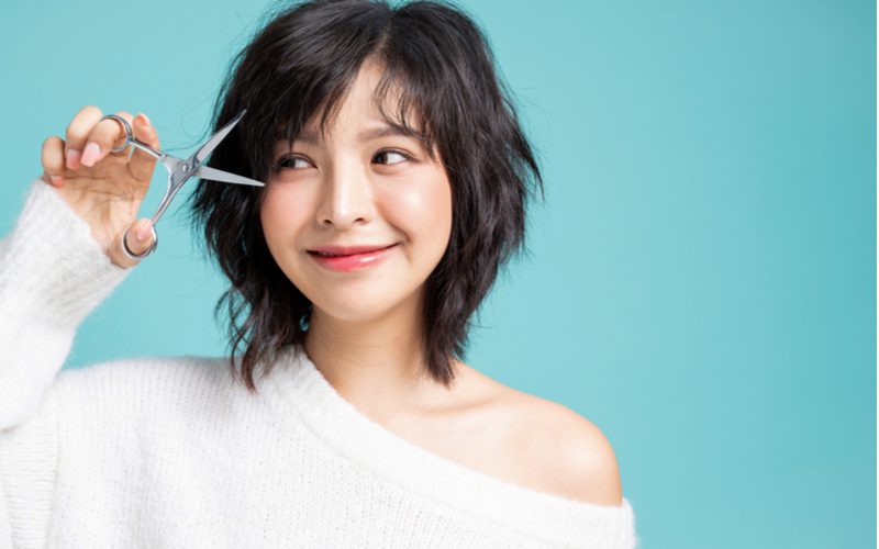 Asian woman in a shoulderless sweater holding up a pair of scissors to symbolize things to consider before cutting bangs