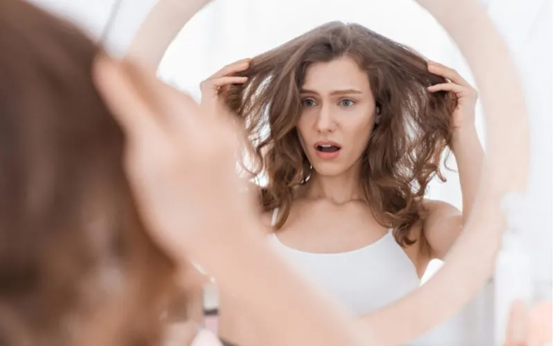 For a piece on how to get rid of greasy hair, a woman holds her hair out and looks in a mirror with a shocked expression on her face