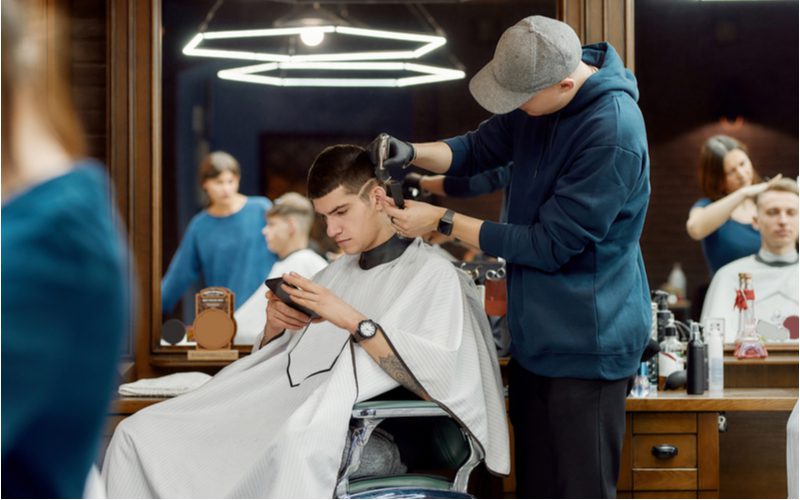 Guy in a barber's chair getting a mid fade haircut from a barber in a dark blue hoodie and a hat