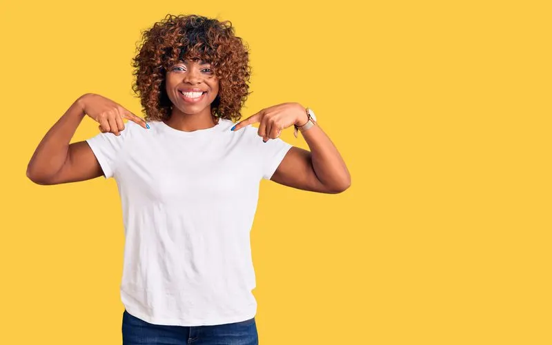 Black woman with a balayage shag haircut stands in a white shirt and points to her chest in a yellow room
