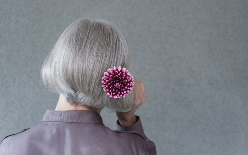 Woman holding a purple flower up to the back of her head in a grey room