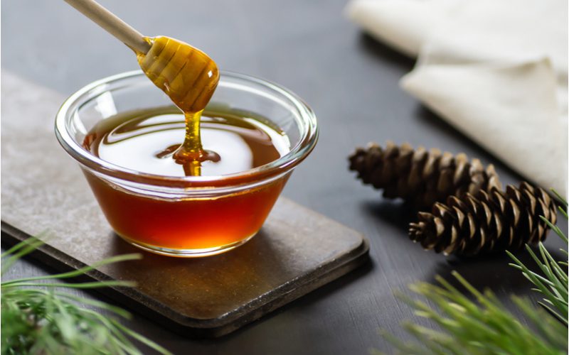 To symbolize a way to lighten hair without bleach, a bowl of honey sits on a table next to pine cones and pine needles