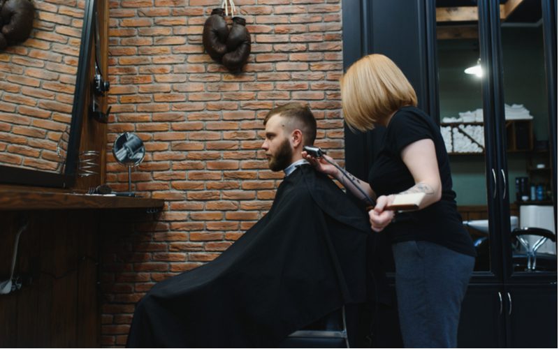 Female barber giving a guy a mohawk fade haircut while he wears a black cape
