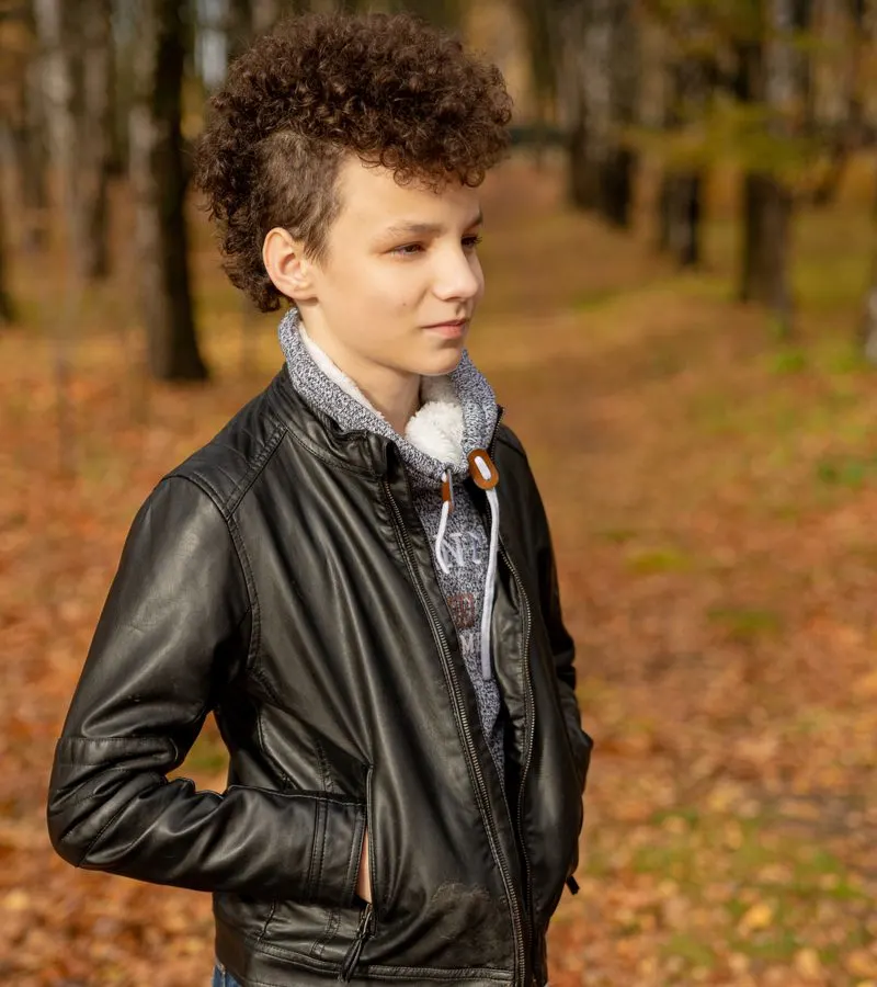 Guy in a leather jacket wearing a Curly Mohawk Mullet Fade while standing in a forest in the Fall