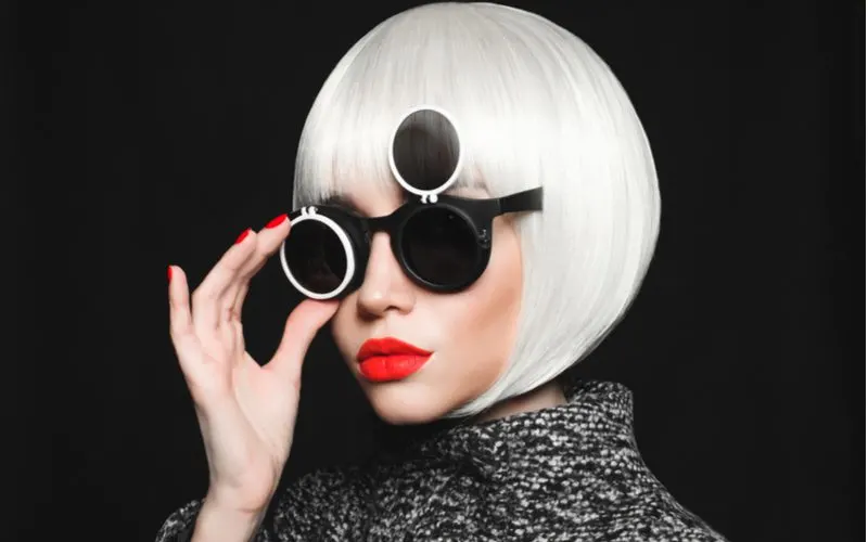 Trendy woman with platinum blonde hair wearing an undercut bob wig and Willie Wonka glasses in a dark room with red lips