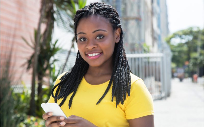Half-Updo Box Braids on a woman in a yellow shirt standing outside a college campus holding a phone