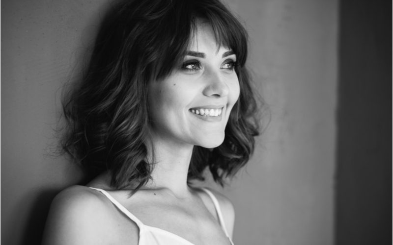 Thin woman with small breasts growing out bangs smiling in a black and white image standing against a wall
