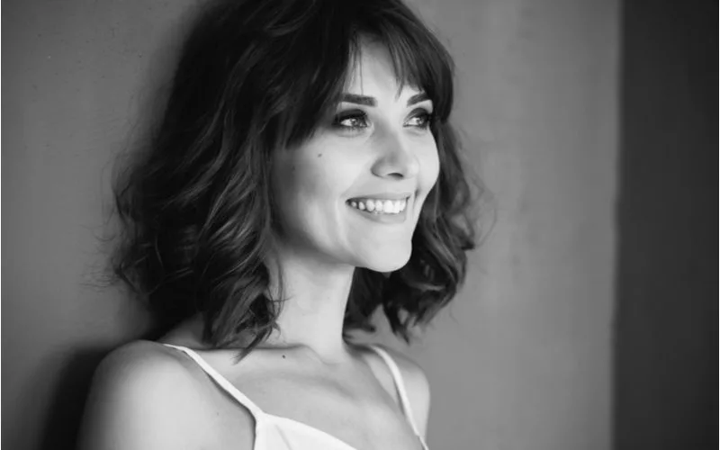 Thin woman with small breasts growing out bangs smiling in a black and white image standing against a wall