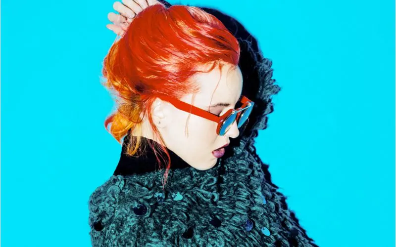 Orange crush punk haircut on a woman in orange glasses standing in a furry teal jacket in a blue room