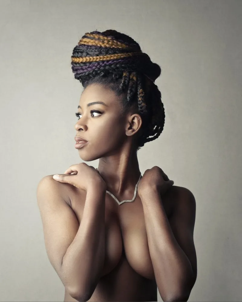 Naked black woman with tribal braids tied into a bun above her head holds her arms in front of her large breasts