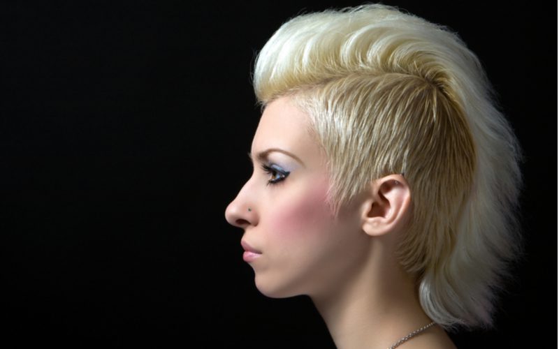 Punk hairstyle titled Shaggy Blonde Mohawk on a woman with pale skin in a black room