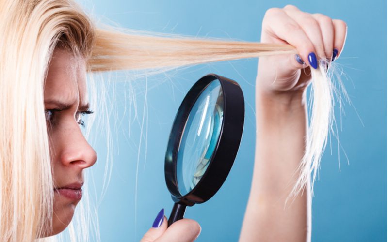 Photo of a woman with a magnifying glass looking at the end of her hair because she bleached it too much in a blue room
