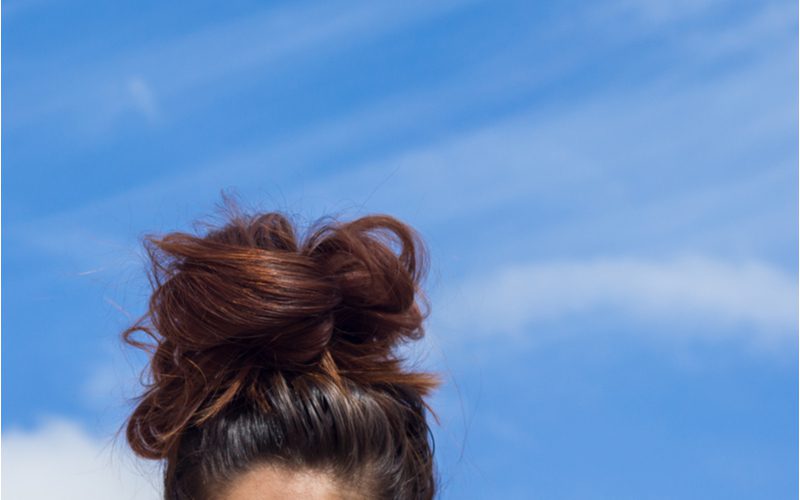 Brunette girl with a messy bun on her head against a blue sky with white clouds for a piece on how to make a messy bun with long hair