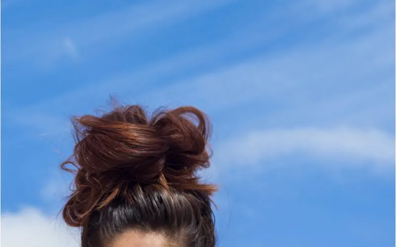 Brunette girl with a messy bun on her head against a blue sky with white clouds for a piece on how to make a messy bun with long hair