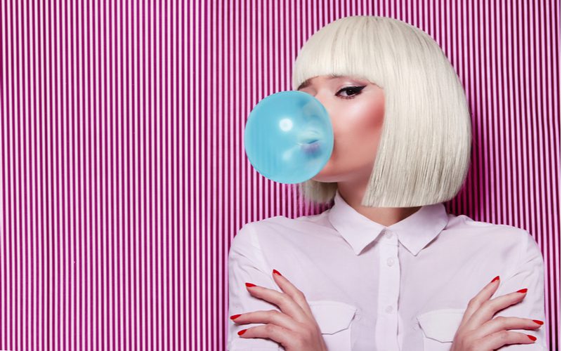 White woman blowing a bubble while wearing a blonde wig and crossing her arms while she grows out her pixie cut