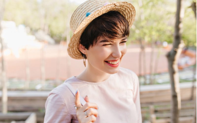 A hack for step 4 in how to grow out a pixie cut shows a woman wearing a hat and smiling while her hair grows