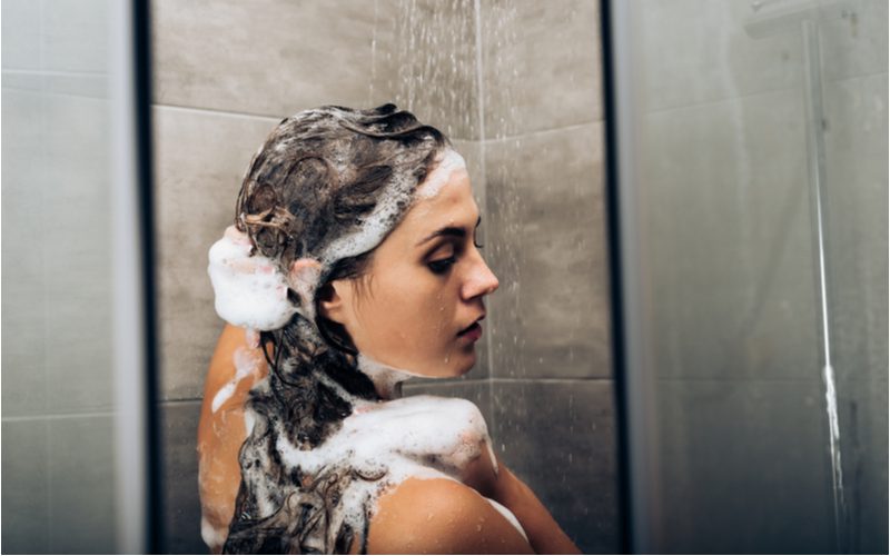 Girl in a tiled shower soaping her long hair while looking over her right shoulder