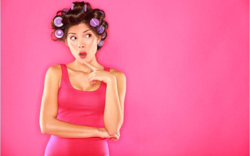 Woman in red lip wearing a red dress in front of a pink wall wondering how to use hair rollers