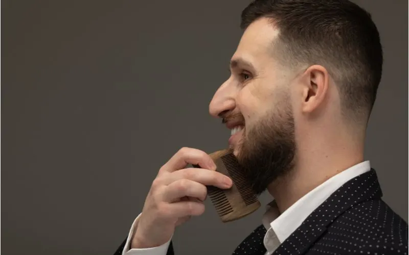 Low Skin Fade With Shaped Edges worn by a smiling bearded man combing his facial hair with a wooden comb