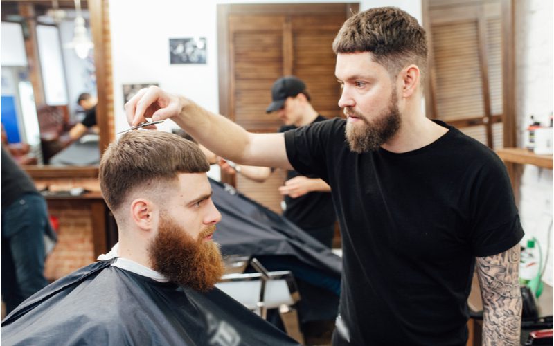 Guy with a Caesar-inspired drop fade and a big bushy beard looking ahead while getting his hair trimmed