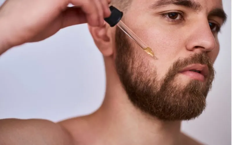 Full short beard on a shirtless man in a studio putting beard oil on his cheek for a piece on styles of beards