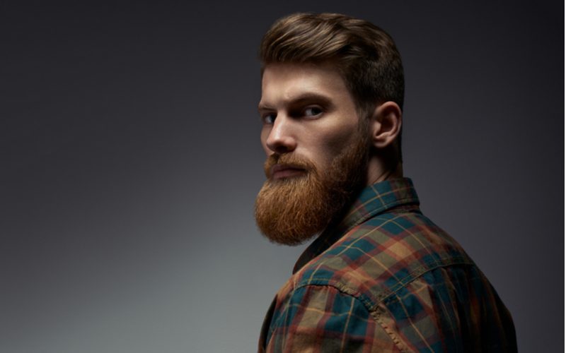 Image for a piece titled Beard Styles featuring a guy rocking a Medium Garibaldi Beard and wearing a red and black flannel shirt standing in a grey, dimly-lit studio