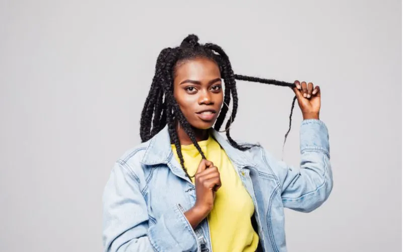 For a piece on dreadlocks, a three strand braided loc style on a woman holding her hair and wearing a yellow shirt with a jean jacket