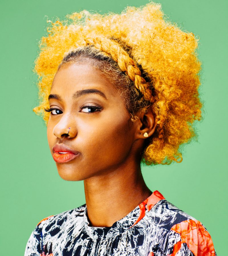 Woman with yellow dyed hair coloring and dark skin wears a button-up shirt with a traditional African pattern on it