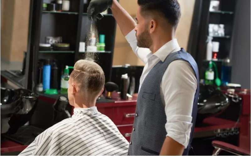 Barber in a vest and long sleeve white button up shirt sprinkling something onto a guy's hair after giving him a low skin fade haircut
