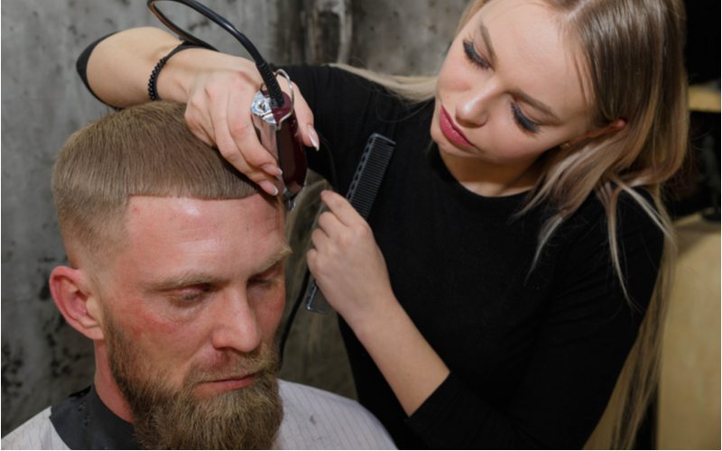 Man in a salon chair gets a Lined-Up Low Bald Caesar Fade haircut from a female hairstylist