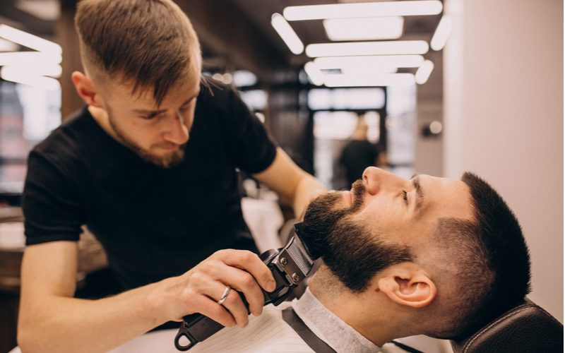 Guy with a low skin fade getting his beard trimmed by a barber while lying back in a barber chair
